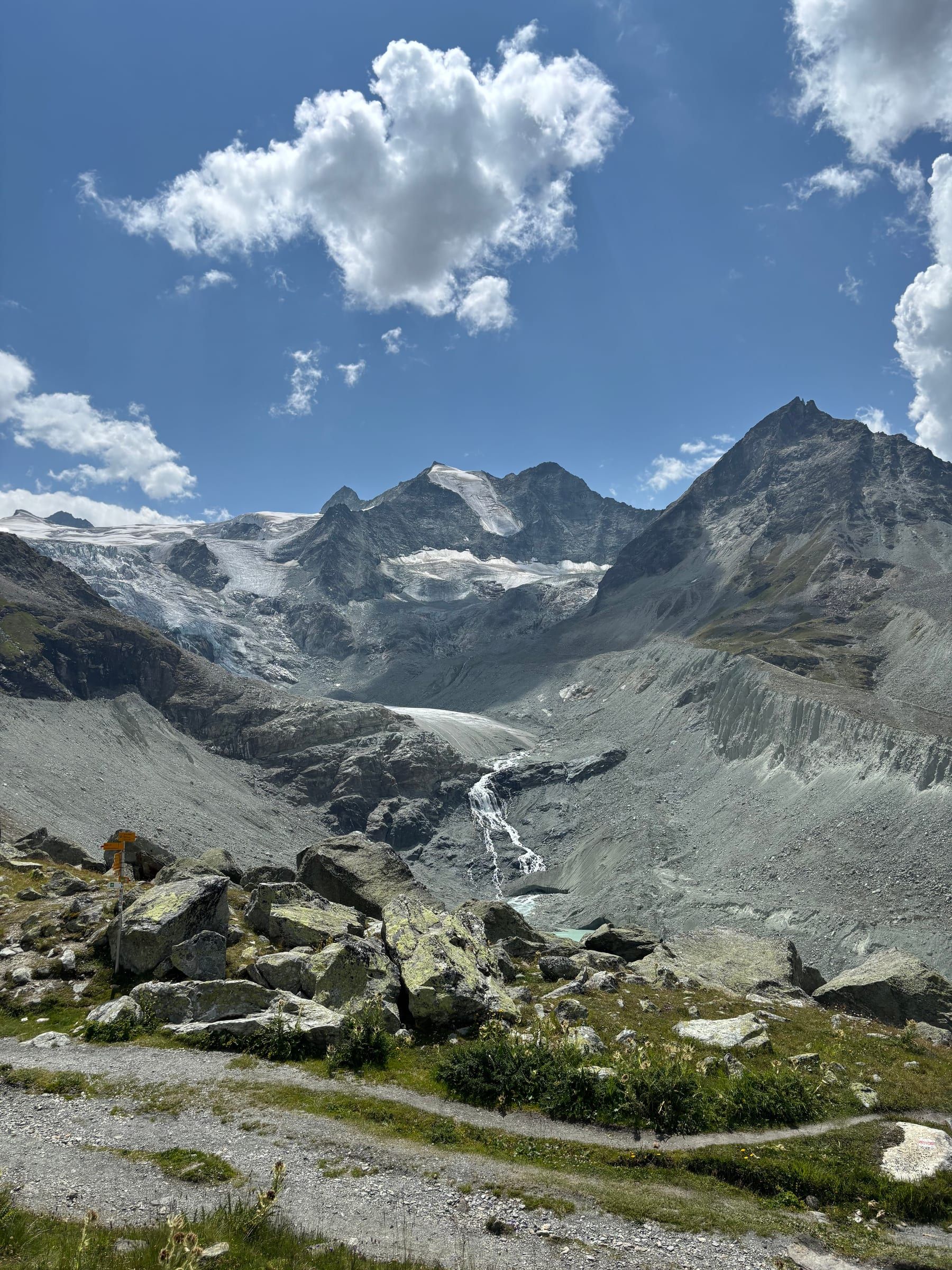 The Moiry glacier and its peaks under a blue sky with clouds, a meltwater waterfall and the rocky trail in the foreground